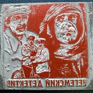 Puede incluir: Placa metálica vintage con un diseño rojo y plateado. La placa presenta imágenes en relieve de varios hombres, incluido un hombre con sombrero y otro con un tocado. El texto "TELEWCKNN VELEKINE" está en la parte inferior.