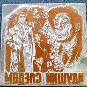 Puede incluir: Placa metálica rectangular con diseño en relieve. El diseño presenta dos hombres, uno con abrigo, y un retrato de un rostro masculino. El texto "IN CUEEDOM" está en la parte inferior. El fondo es de color naranja óxido.