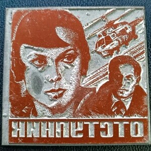 Puede incluir: Pin metálico cuadrado, rojo y plateado, con dos retratos y una nave espacial. La mitad superior del pin tiene un retrato de una mujer y una nave espacial. La mitad inferior tiene un retrato de un hombre y texto en un idioma extranjero.