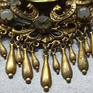 May include: Close-up of a vintage gold-toned brooch with intricate filigree details. The brooch features teardrop-shaped dangles and small, round, off-white stones. The design is ornate and detailed, with a classic, antique aesthetic.