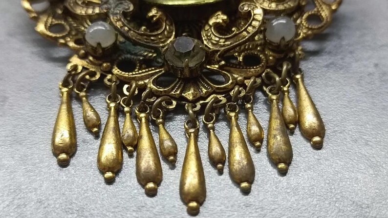 May include: Close-up of a vintage gold-toned brooch with intricate filigree details. The brooch features teardrop-shaped dangles and small, round, off-white stones. The design is ornate and detailed, with a classic, antique aesthetic.