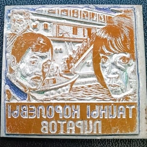 Puede incluir: Una impresión en relieve rectangular de color cobre con dos caras, un barco y texto. Las caras están detalladas con emoción visible. El barco está en el fondo. El texto está en un idioma extranjero. La impresión tiene un borde azul y blanco.