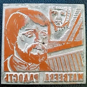Puede incluir: Un bloque de impresión metálico cuadrado con un diseño naranja y plateado. El diseño presenta un retrato de un hombre barbudo y un segundo rostro en el fondo. El texto "NILBERT RAAVOGIP" está impreso al revés en la parte inferior del bloque.