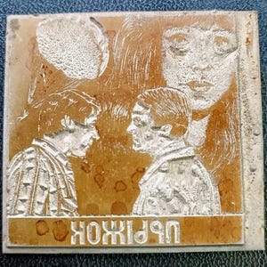 Puede incluir: Placa cuadrada de metal envejecido con un diseño grabado. El diseño presenta a dos hombres enfrentados, un rostro de mujer y texto cirílico en la parte inferior. El metal tiene una combinación de colores marrón y plateado.
