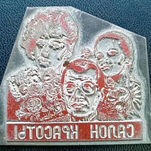 Puede incluir: Un bloque de impresión metálico con tres caras y detalles en rojo. Diseños florales rodean las caras. El texto "HONAS KBCOLPI" está impreso en rojo en la parte inferior del bloque.