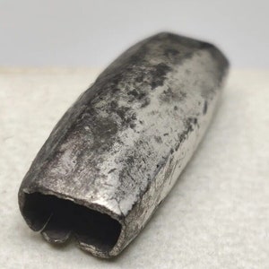 Puede incluir: Un tubo de metal plateado con una forma ligeramente redondeada y una abertura rectangular en un extremo. El tubo tiene un aspecto desgastado y envejecido.