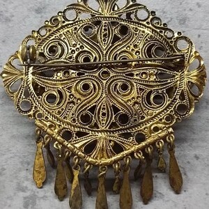 May include: An antique gold-coloured brooch with an oval filigree design. The brooch has detailed patterns, openwork, and dangling teardrop charms. A classic piece of jewellery.