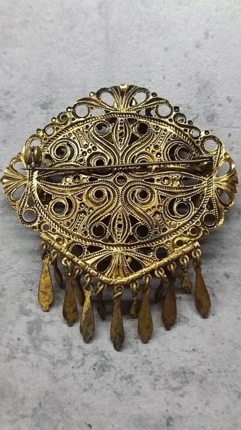 May include: An antique gold-coloured brooch with an oval filigree design. The brooch has detailed patterns, openwork, and dangling teardrop charms. A classic piece of jewellery.