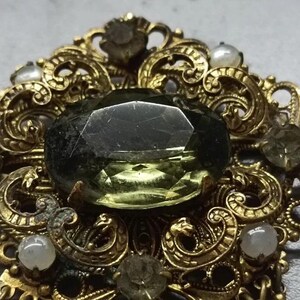 May include: An ornate gold brooch with a large, faceted, olive-green central stone. The brooch features intricate filigree detailing and is accented with smaller, round, off-white and olive-green stones. A vintage-style jewellery piece.