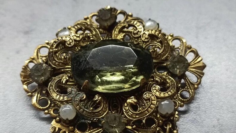 May include: An ornate gold brooch with a large, faceted, olive-green central stone. The brooch features intricate filigree detailing and is accented with smaller, round, off-white and olive-green stones. A vintage-style jewellery piece.