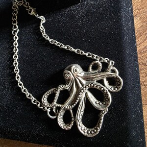 May include: Silver-toned octopus pendant necklace. The pendant features a detailed octopus design with textured tentacles and a chain. The necklace is displayed on a black surface, showcasing the intricate details of the jewelry.