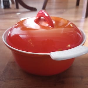 May include: A red and white enameled cast iron pot with a lid and a white handle. The pot is sitting on a wooden surface.
