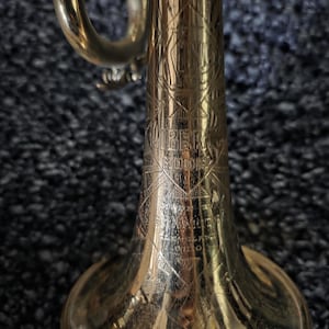 May include: A close-up of a brass trumpet with intricate engraved details. The trumpet has a bell shape and a curved mouthpiece. The text "Liberty Model" is visible on the body of the instrument. The trumpet is a musical instrument.