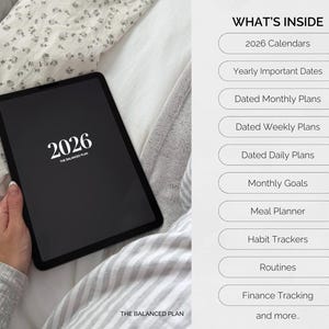 May include: A black tablet displaying the year 2026 in white text, with the words "THE BALANCED PLAN" below. The image also lists features such as calendars, monthly plans, and habit trackers. The tablet is held on a bed with a floral patterned sheet.
