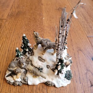 May include: A ceramic figurine of a wolf family in a snowy forest setting. The figurine features two wolves howling at the moon, a smaller wolf pup, and a bird. The base of the figurine is decorated with snow, trees, and footprints.