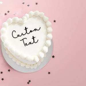 May include: A white heart-shaped cake with white frosting and the words "Custom Text" written in black on top. The cake is on a white plate and surrounded by silver stars on a pink background.