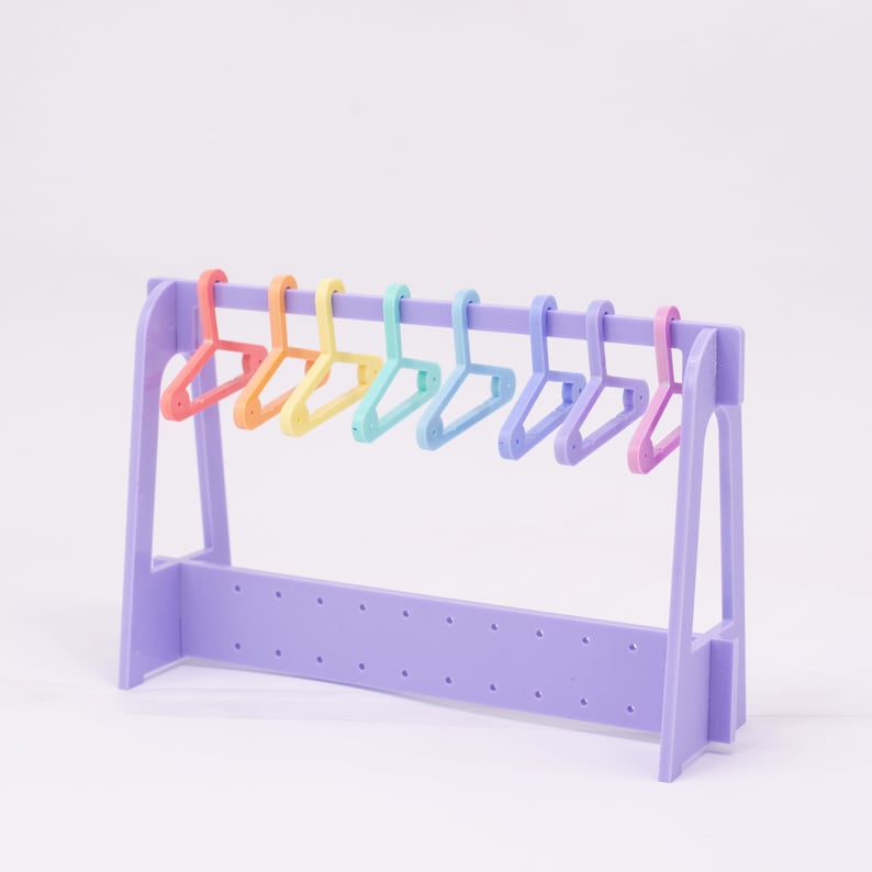 Puede incluir: Un perchero miniatura de color morado con ocho perchas de pl&aacute;stico de colores arco&iacute;ris.