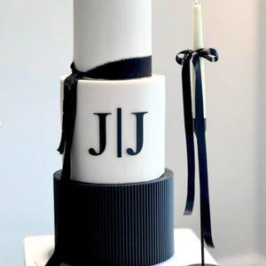 May include: A three-tier wedding cake decorated with black and white fondant. The top tier is white with a black ribbon and the middle tier is white with the initials "JIJ" in black. The bottom tier is black with a ribbed design.