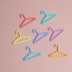Puede incluir: Siete perchas de pl&aacute;stico de colores pastel, incluyendo rosa, azul, amarillo y verde. Las perchas est&aacute;n dispuestas sobre un fondo rosa.