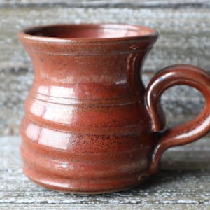 May include: A handmade ceramic mug with a reddish-brown glaze. The mug has a rounded body and a wide, flared rim. The handle is attached to the side of the mug and curves up and over the top.