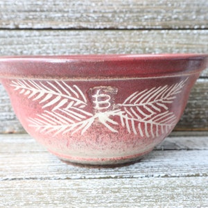 May include: A maroon ceramic mixing bowl with a white leaf design. The bowl has a wide rim and a slightly textured surface. The leaf design is repeated around the bowl, with a letter "B" in the center.