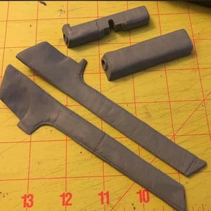 May include: Grey automotive interior trim components, including two long, angled pieces, and several smaller parts. The items are arranged on a yellow cutting mat with black grid lines and number markings.