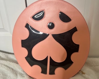 May include: A round, pink leather bag with a black, heart-shaped design. The bag has a zipper closure and a gold zipper pull.