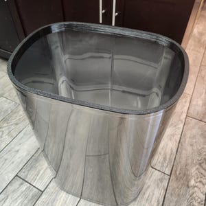 May include: A tall, oval-shaped trash can with a polished, reflective stainless steel exterior. The top rim features a thick, textured black border that contrasts with the smooth, metallic body. The interior is deep and finished in a dark, glossy tone.