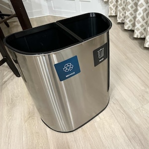 May include: A stainless steel dual compartment trash and recycling bin. The bin has a black interior and a blue label that says "RECYCLE" with a recycling symbol. A black label on the bin says "TRASH".