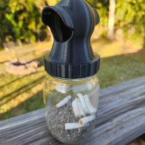 Peut inclure: Un couvercle de bocal en plastique noir avec un bec verseur pour éteindre les cigarettes. Le bocal est rempli de mégots de cigarettes et de sable.