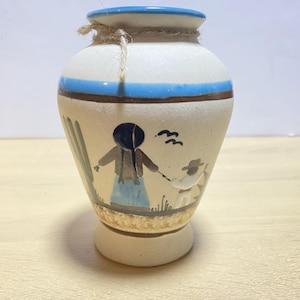 Könnte beinhalten: Eine Keramikvase mit cremefarbenem Körper, verziert mit einem blauen Band am oberen Rand und einem braunen Band darunter. Die Vase zeigt eine handgemalte Szene mit Figuren und einem Kaktus. Um den Hals ist eine Schnur aus Bindfaden gebunden.