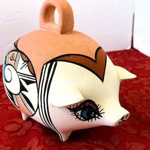 May include: A ceramic piggy bank with a peach-colored top and a white body. The piggy bank features a decorative design with black, brown, and blue accents. It has a loop handle and a whimsical eye design.