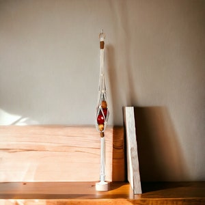 May include: A white macrame hanging planter with a wooden ring at the top. The planter is filled with red and orange fruit. The planter is hanging from a wooden shelf.