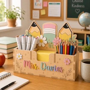 May include: A wooden desk organiser featuring a pencil and eraser design with floral accents. The front is personalised with the text "Mrs. Davies" in colourful script. It includes compartments holding markers, coloured pencils, and a yellow sticky note pad.