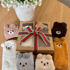 May include: A gift box tied with a red, green, and gold striped ribbon, surrounded by six pairs of fuzzy, plush socks in white, gray, tan, brown, and mustard yellow, each featuring a small embroidered bear face with protruding ears.