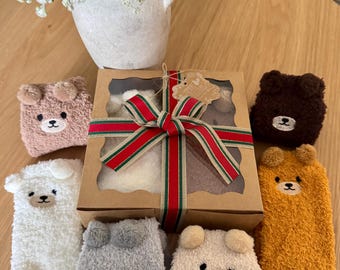 May include: A gift box tied with a red, green, and gold striped ribbon, surrounded by six pairs of fuzzy, plush socks in white, gray, tan, brown, and mustard yellow, each featuring a small embroidered bear face with protruding ears.