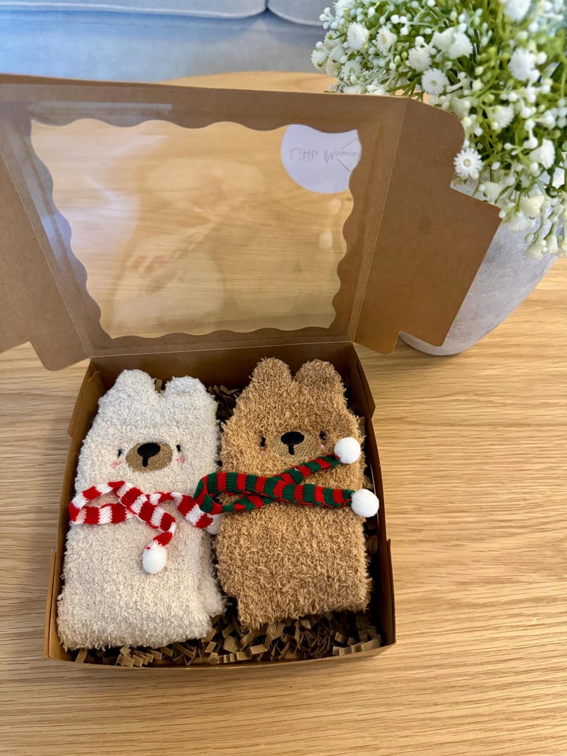 May include: A gift box containing two fuzzy bear-shaped socks. One sock is cream-colored, the other is brown. Each sock has a face and is adorned with a red and white or green and red striped scarf. The box has a clear window.