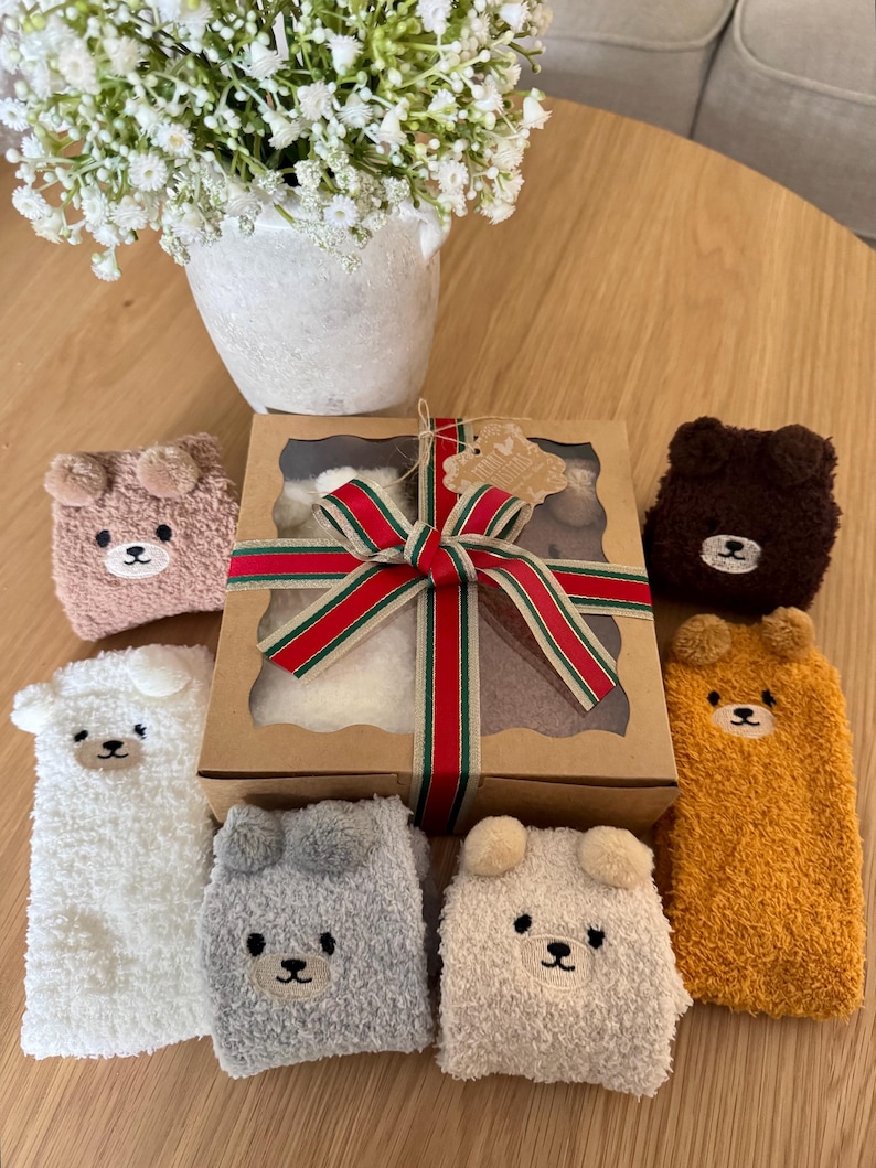 May include: A gift box tied with a red, green, and gold striped ribbon, surrounded by six pairs of fuzzy, plush socks in white, gray, tan, brown, and mustard yellow, each featuring a small embroidered bear face with protruding ears.