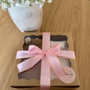 May include: A square cardboard gift box with a clear window, tied with a pink satin ribbon. Inside, brown and white fuzzy items are visible. A small round sticker with the text "Moments" in a diamond-shaped frame is attached to the box corner.