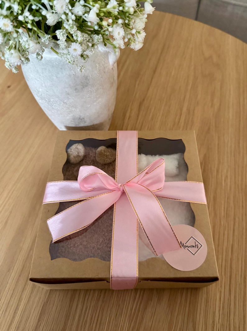 May include: A square cardboard gift box with a clear window, tied with a pink satin ribbon. Inside, brown and white fuzzy items are visible. A small round sticker with the text "Moments" in a diamond-shaped frame is attached to the box corner.