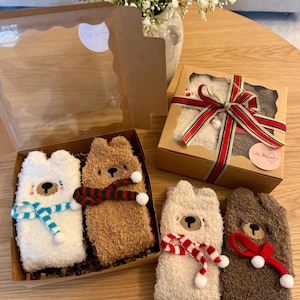 May include: Four pairs of bear-shaped socks in gift boxes. The socks are white, beige, and brown, with embroidered faces and scarves in various colours. One box is tied with a red and white striped ribbon. The socks are displayed on a wooden table.