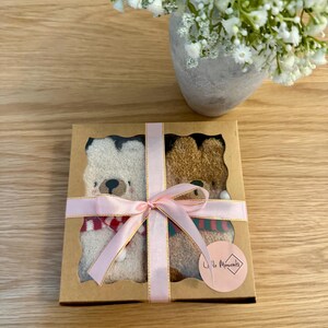 May include: A gift box with two teddy bear socks, one cream and one brown, each with a scarf. The box is tied with a pink ribbon. A vase of white flowers is in the background.