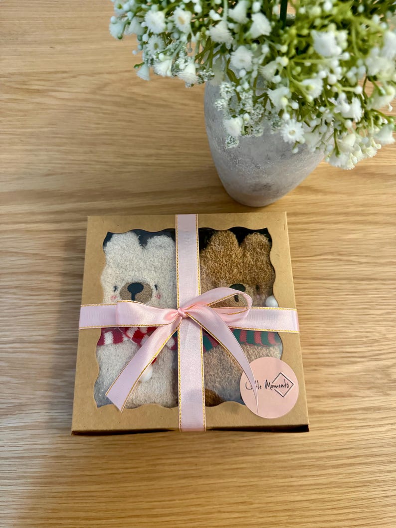May include: A gift box with two teddy bear socks, one cream and one brown, each with a scarf. The box is tied with a pink ribbon. A vase of white flowers is in the background.