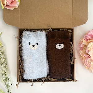 May include: A pair of fuzzy socks in a cardboard gift box, one light gray and one dark brown, each featuring a stitched bear face with small ears at the top. The socks are nestled in brown paper filler, presented alongside pink and white flowers.