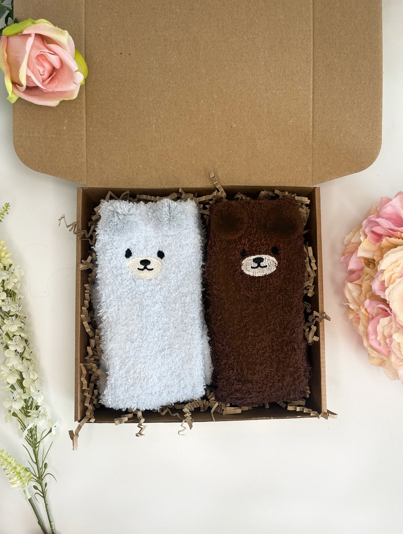 May include: A pair of fuzzy socks in a cardboard gift box, one light gray and one dark brown, each featuring a stitched bear face with small ears at the top. The socks are nestled in brown paper filler, presented alongside pink and white flowers.