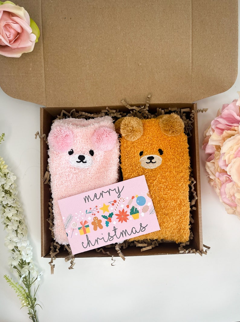 May include: A pair of fuzzy plush socks, one light pink and one golden brown, each featuring bear ears and a bear face, nestled in a cardboard gift box with brown paper filler and a pink card printed with the text merry christmas.