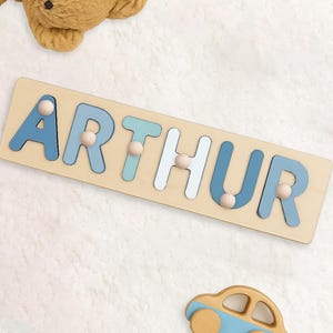 May include: Wooden puzzle spelling "ARTHUR" in blue and white letters with wooden pegs. A small wooden car with blue accents sits below the puzzle. A plush teddy bear is in the background.