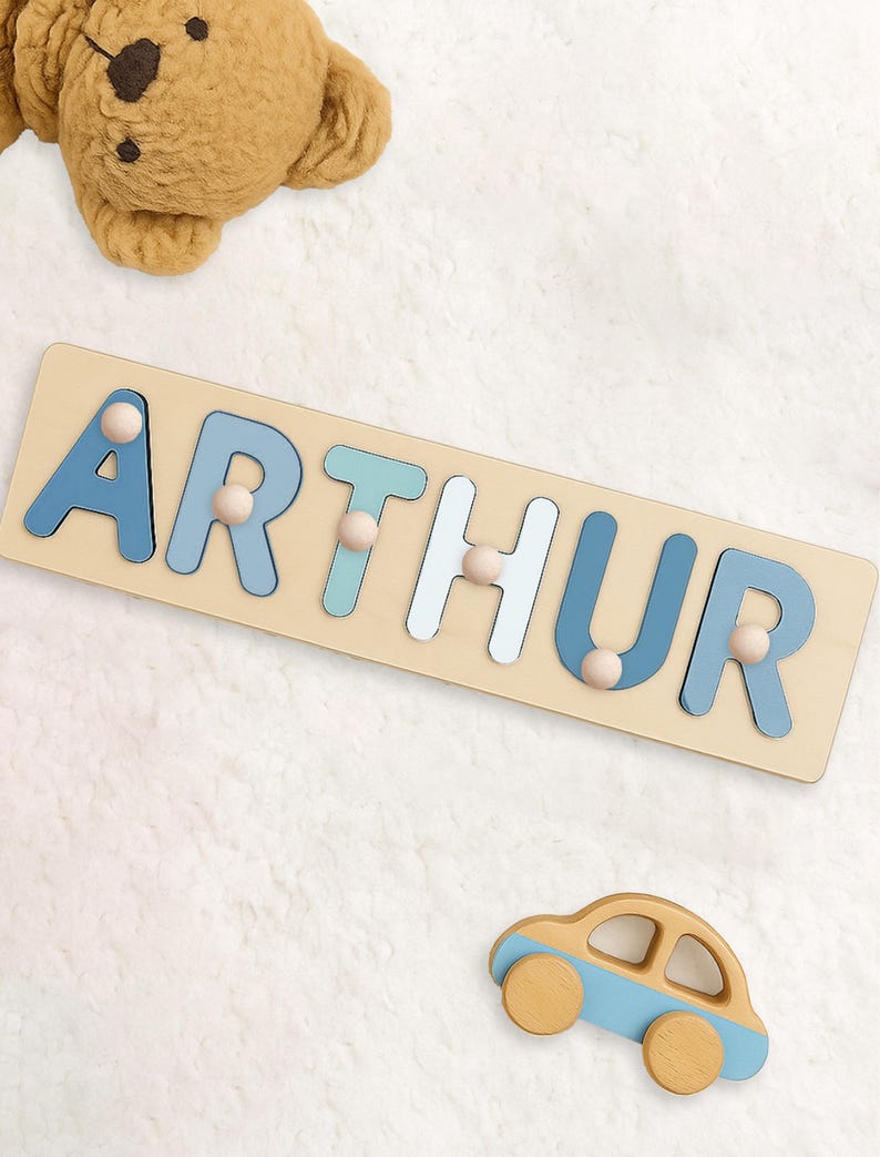 May include: Wooden puzzle spelling "ARTHUR" in blue and white letters with wooden pegs. A small wooden car with blue accents sits below the puzzle. A plush teddy bear is in the background.