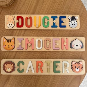 May include: Three wooden name puzzles with colorful letters and animal-themed end pieces. The names are "Dougie", "Imogen", and "Carter". Each puzzle has a giraffe, zebra, cat, dog, lion, and tiger design. The puzzles are on a light brown wooden surface.