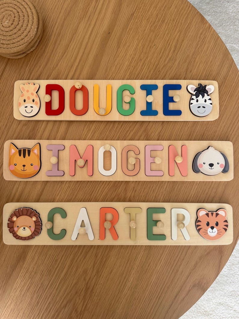 May include: Three wooden name puzzles with colorful letters and animal-themed end pieces. The names are "Dougie", "Imogen", and "Carter". Each puzzle has a giraffe, zebra, cat, dog, lion, and tiger design. The puzzles are on a light brown wooden surface.